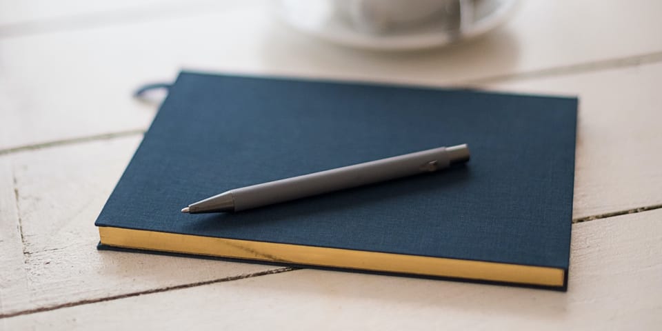 A pen and a closed notebook on a table, next to a cup of coffee.